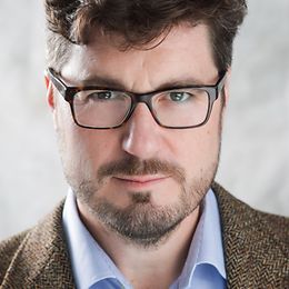Close-up of a man with a beard, glasses and a tweed jacket without a tie, looking friendly into the camera.