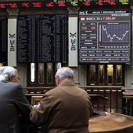 Zwei Händler diskutieren vor einem Display mit Börsenkursen 