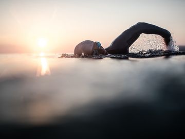A person wearing a neoprene suit, swimming cap and goggles is swimming in open water at sunrise