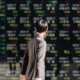 A man in the street looks at a screen displaying stock market prices as he passes.