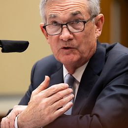 Close-up of a grey-haired man wearing glasses, tie and suit, gesticulating as he speaks into a microphone.