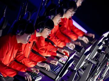 Vier junge Männer mit identischen roten Jacken, Kopfhörern und Konsolen starren konzentriert auf ihre Bildschirme