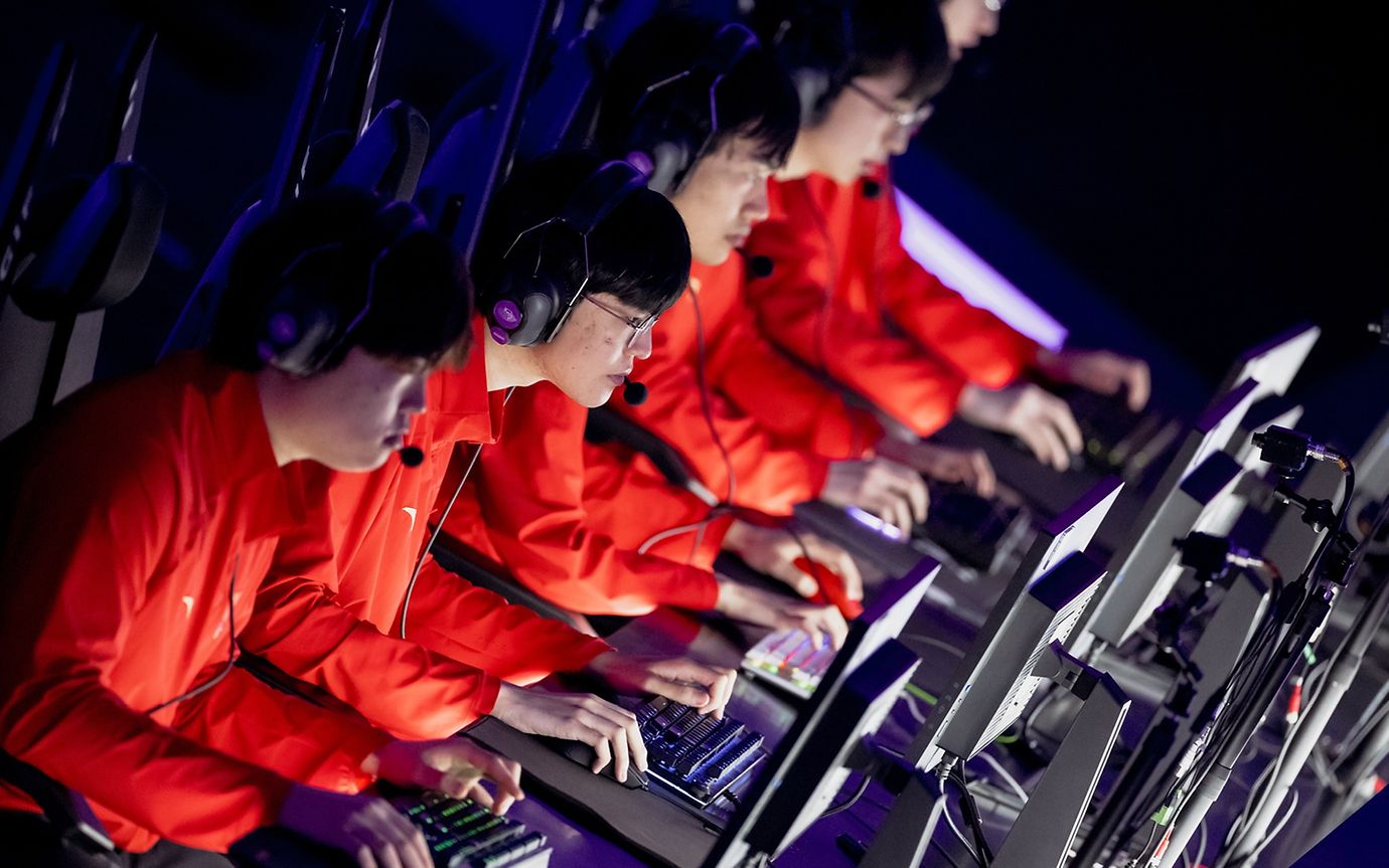 Vier junge Männer mit identischen roten Jacken, Kopfhörern und Konsolen starren konzentriert auf ihre Bildschirme