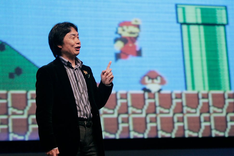 Ein Mann spricht vor einem grossen Bildschirm, auf dem das Videospiel Mario Brothers läuft