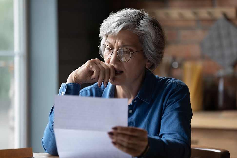 A bespectacled woman is sitting in the kitchen, looking thoughtfully, perhaps anxiously, at a document.