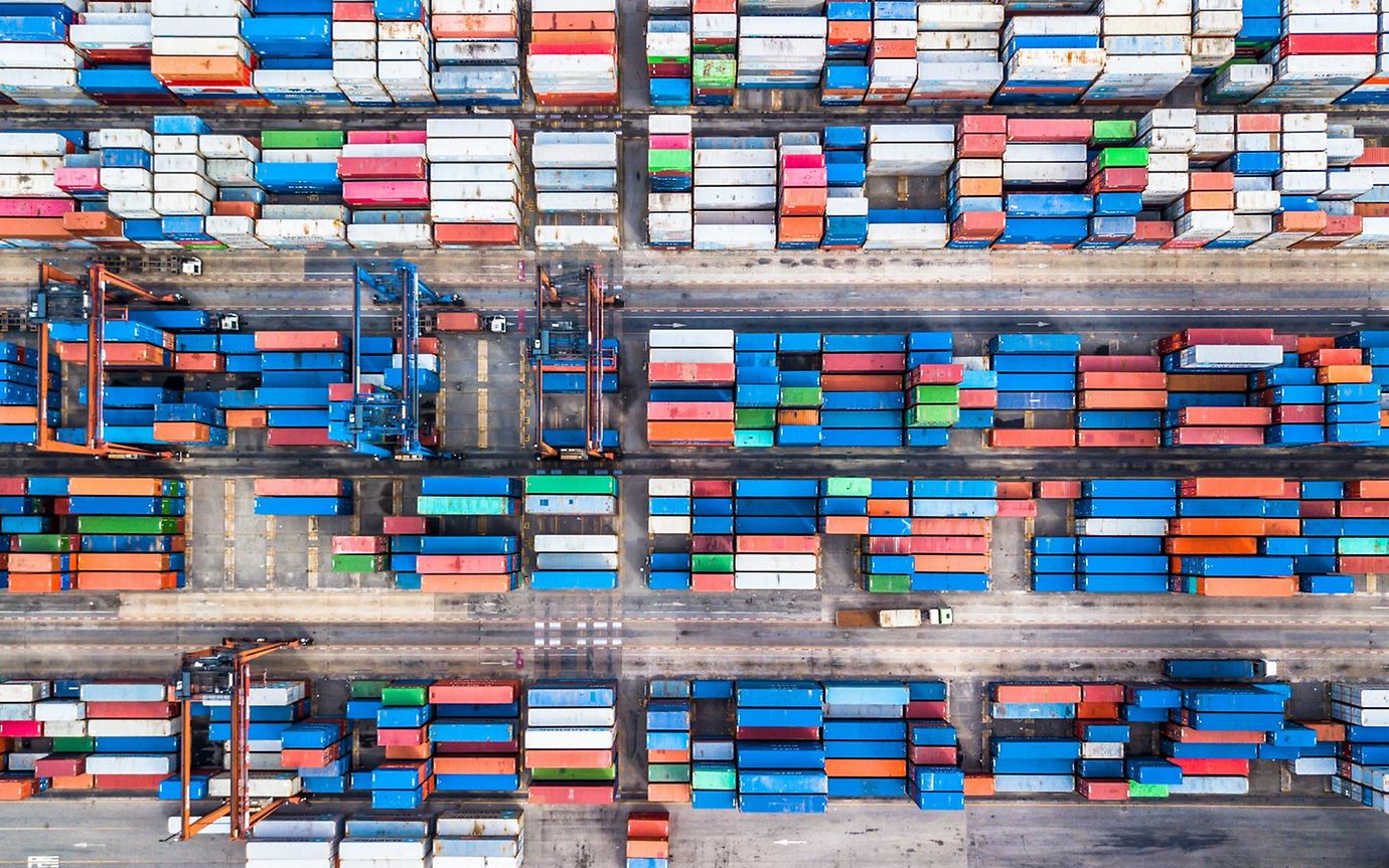 Viele verschiedenfarbige Container in einem Containerhafen, aus der Vogelperspektive sieht es wie ein Mosaik aus.