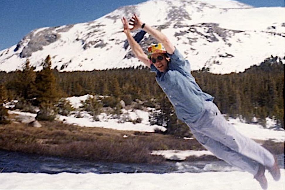 A casually dressed man wearing a sun hat and sunglasses jumps into the snow against a mountain backdrop.