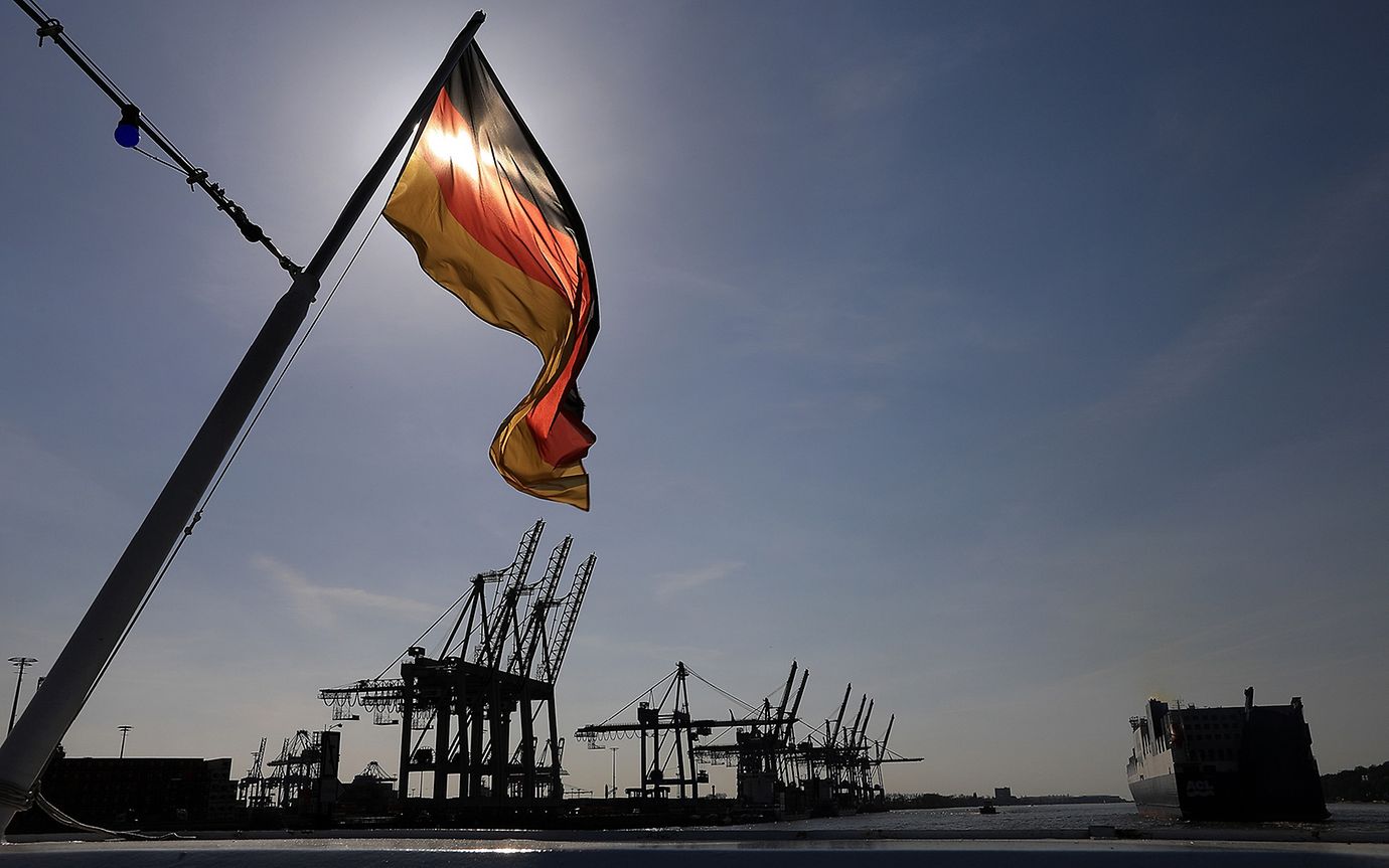 A German flag flies, behind it, ship cranes stand in a German harbour at dusk.