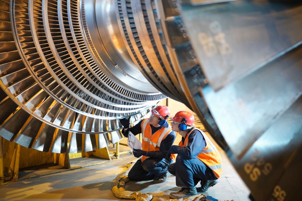 Two people in work clothes and hard hats are inspecting a large turbine