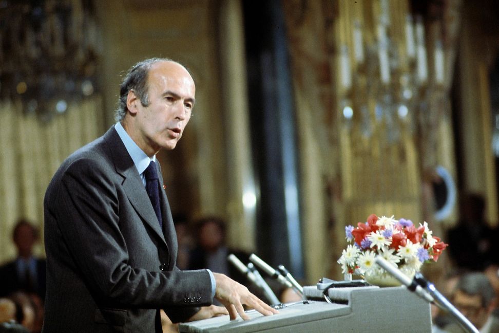 A man in a suit and tie is speaking in a historic government building. The image quality makes it clear that the image is dated.