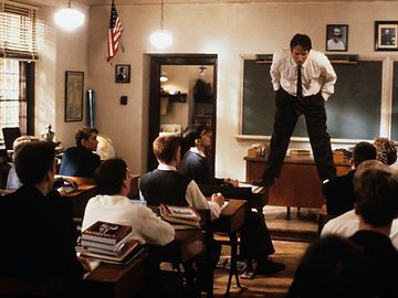 Ein Englischlehrer steht auf einem Pult in einem Klassenzimmer und spricht zu den Schülern, die zu ihm aufschauen.