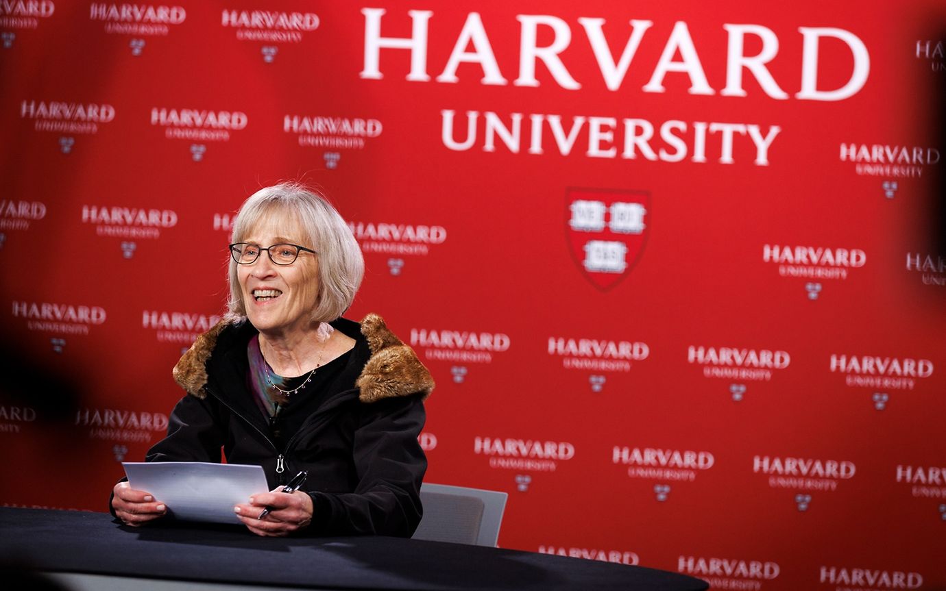 Eine Frau mit Brille und Unterlagen spricht vor einer Wand mit der Aufschrift Harvard University und dem Logo.