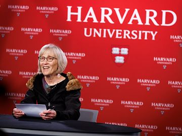 A woman with glasses and documents is speaking in front of a wall with the Harvard University logo.