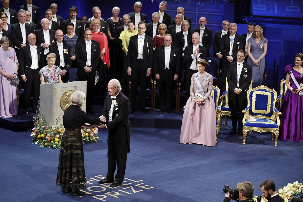 A woman in evening dress accepts a gift from a man at a royal court surrounded by people.