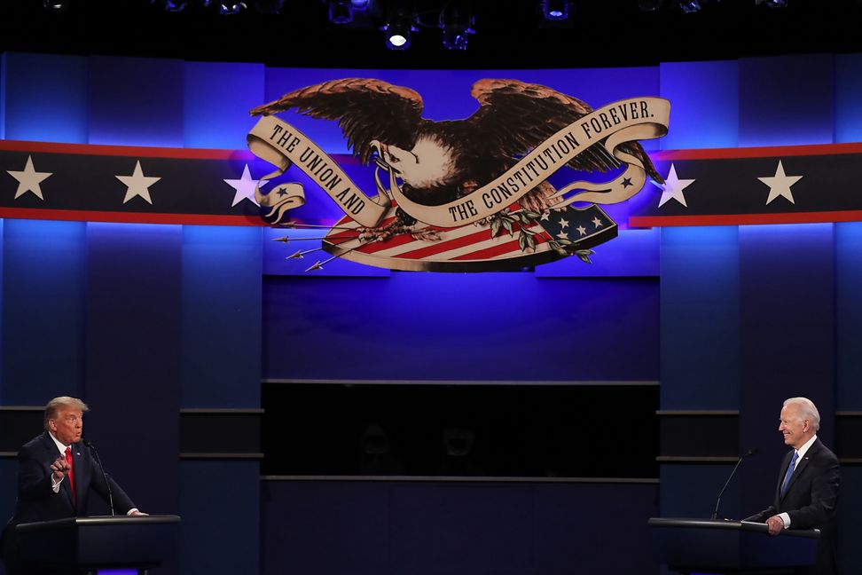 Two people at lecterns in an auditorium under the coat of arms of the USA.