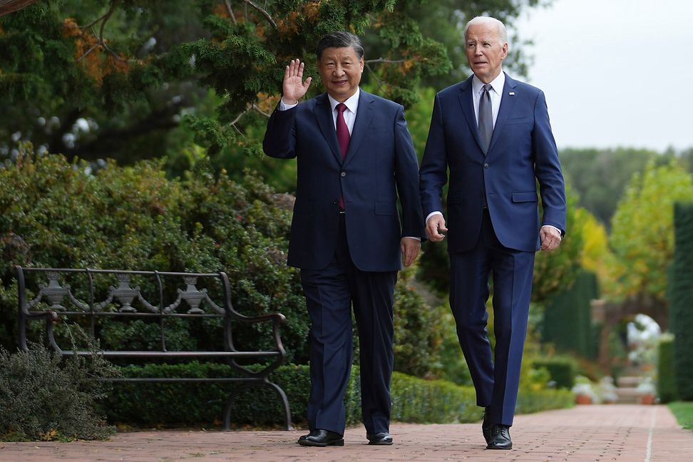 Two men, both wearing suits, are walking through a garden. One waves to the photographers.