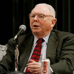 An elderly man with thick glasses, suit and tie, Diet Coke next to him, speaks into a microphone