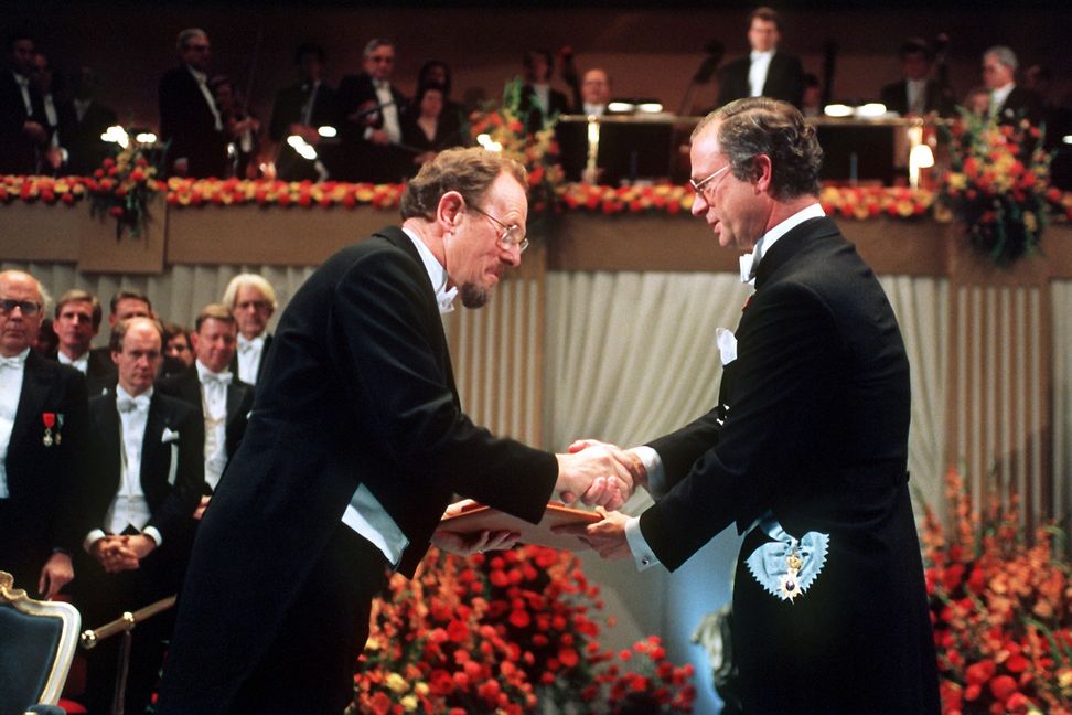 Two men in evening dress shake hands in front of an audience, one bowing slightly to the other
