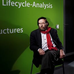 Männlich mittleren Alters mit rotem Schal sitzt mit übergeschlagenen Beinen auf einem Stuhl vor einer grünen Wand.