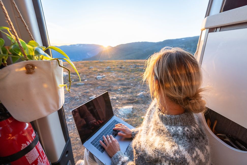 Junge Frau sitzt im Camper mit Blick auf eine einsame Hochebene, den Laptop auf dem Schoss und beobachtet den Sonnenuntergang.