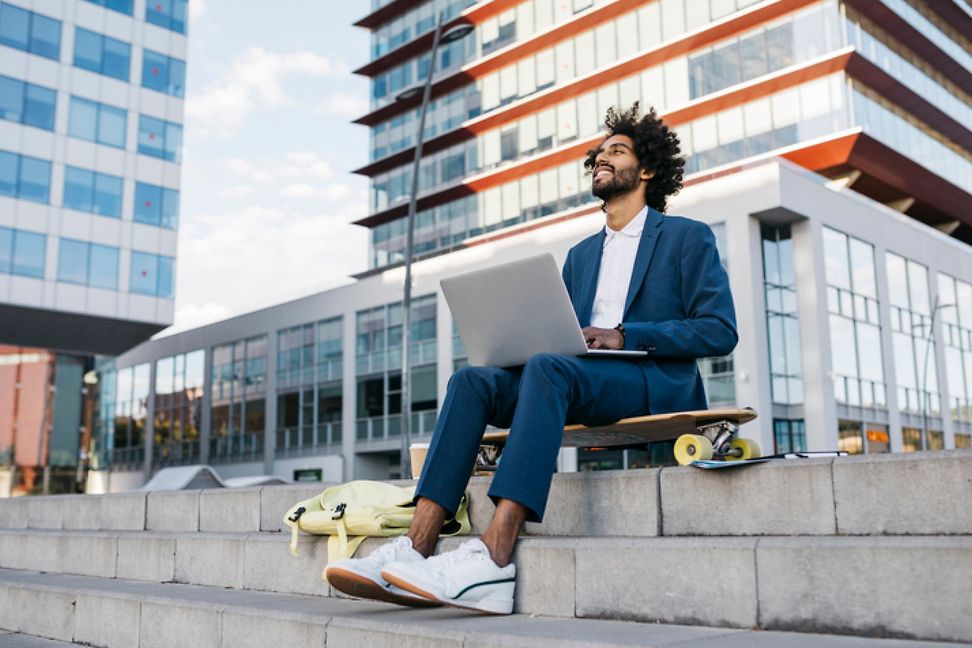 Young man in a suit and sneakers sits on a skateboard with a laptop on his lap in front of a skyline.