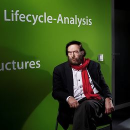 A middle-aged male intellectual wearing a red scarf sits on a chair with his legs crossed in front of a green wall.