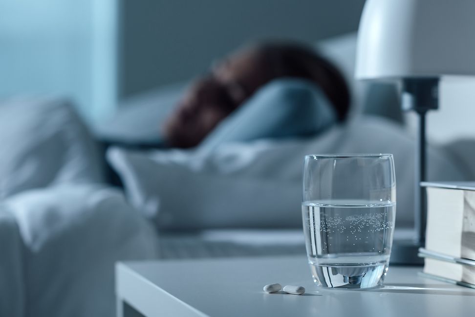 Pillen auf Nachttisch neben Wasserglas neben schlafender Person im Bett