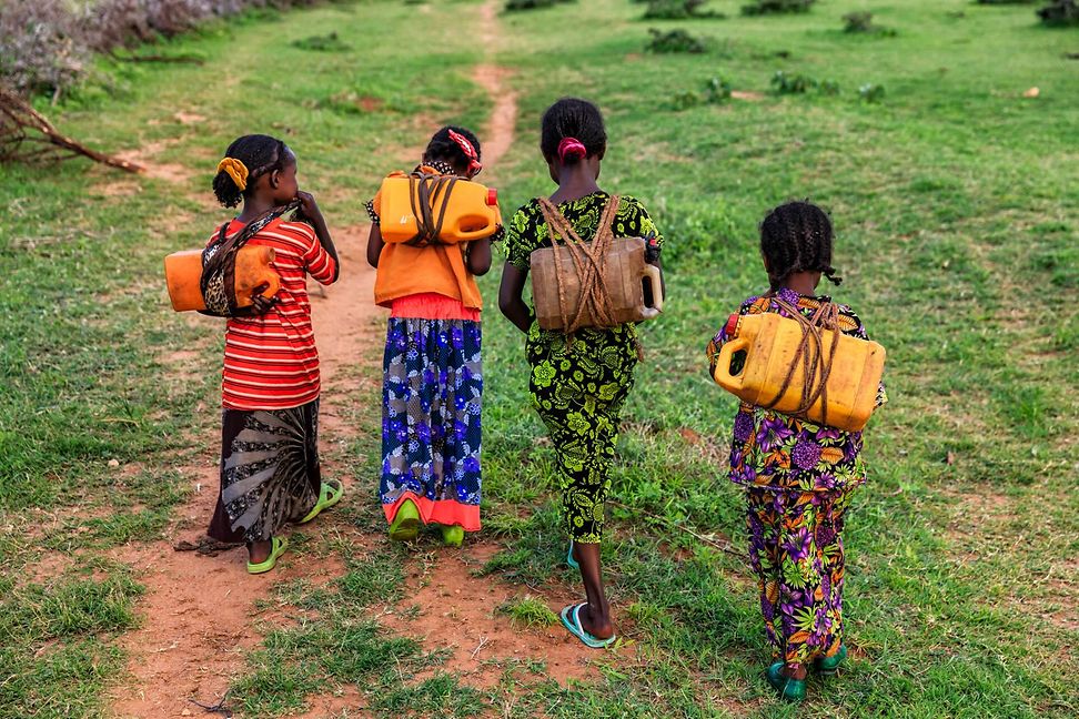 Vier Mädchen in bunten afrikanischen Gewändern tragen Wasserkanister auf dem Rücken.