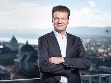 Reto Knutti, auf einer Terrasse stehend mit dem Zürichsee und der Universität im Hintergrund, blickt in die Kamera.