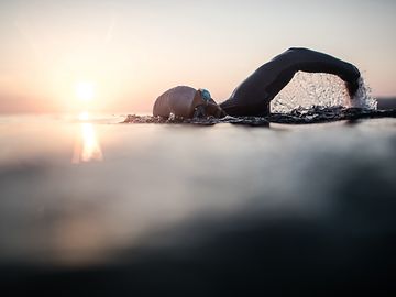 Eine Person, bekleidet mit Neopren, Badekappe und Schwimmbrille, schwimmt bei Sonnenaufgang im offenen Wasser